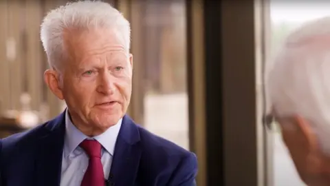 ISHLT An older man with sharp white hair, wearing a navy blue suit, light blue shirt and crimson tie, is being interviewed by another man with white hair, whose head we see the back of.
