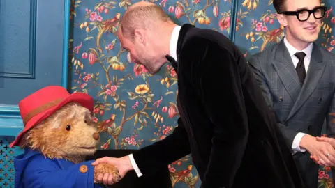 Reuters The Prince of Wales, wearing a black tuxedo, shaking the hand of an actor in costume as Paddington Bear - Tom Fletcher from McFly can be seen in the background 