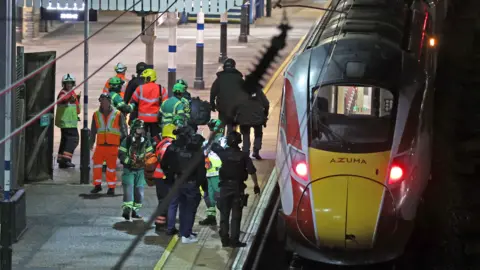 PA Media A group of people in high-vis green and orange vests and jackets crowd on a platform beside a train. It is after dark.