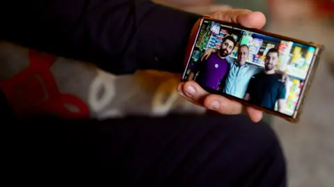 BBC/Goktay Koraltan A hand holds a phone showing of a photo of an older man with his arms round two younger men