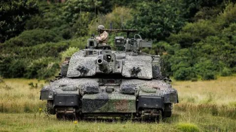Ministry of Defence The Megatron variant of the Challenger 2 tank, heavily camouflaged, with a soldier wearing a helmet in the turret, advancing across a green field, with trees in the background
