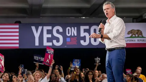 Getty Images California Gov Gavin Newsom appears on stage in support of a redistricting effort in his state. People in the audience are holding up signs in support of Prop 50 and on the wall in the background is a "YES ON 50" sign. 