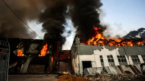 Gallo Images via Getty Images A factory in flames. Bright orange fire and thick black smoke can be seen coming from the top floor of the building. 