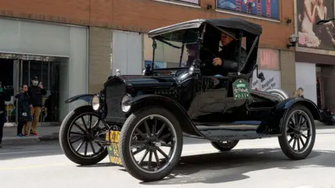 NurPhoto via Getty Images A Ford Model T car is driven on a road. A couple stand on the pavement watching the car go by. One of them is taking a picture of it on their phone. 