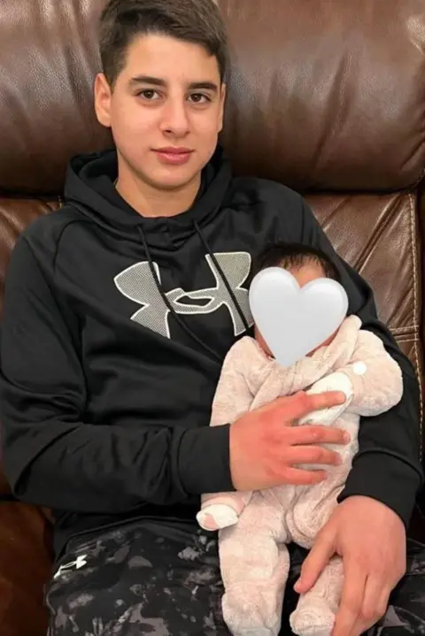 Zaher Ibrahim Mohammed is sitting on a leather sofa and holding a small baby whose face has been covered with a heart emoji. He is smiling and wears a black hooded jumper and patterned trousers, and the baby is in a pink fleece onesie. 