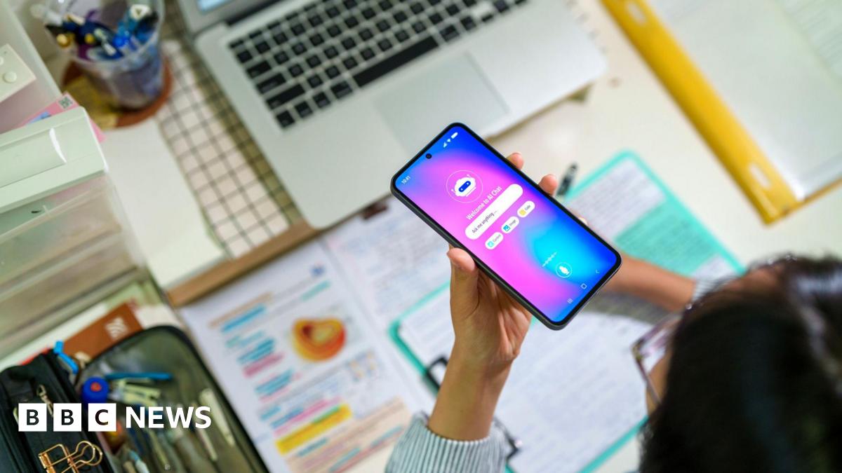 A young female student seen from above interacts with an AI chatbot on a smartphone while studying at a desk with a laptop, notes and stationery. The scene highlights modern learning and technology integration.
