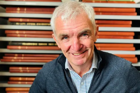 Billy Watson, who has grey hair and is wearing a blue checked shirt and blue top, is smiling at the camera. There are bound copies of old Fraserburgh Herald newspapers behind him.