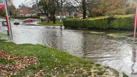 A residential street that has flood water completely covering it. There is a bus stop sign on the left, and grass next to it. There is a black car driving towards the water in the distance