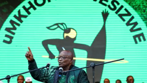 Reuters Jacob Zuma is addressing a rally, with a logo of his party - a spear and a shield - in the background