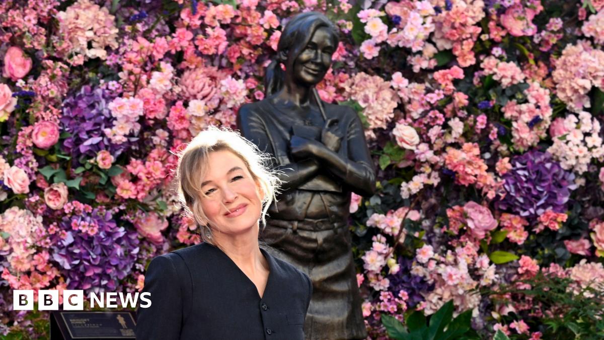 Renée Zellweger smiles while wearing a black dress and posing next to a statue of Bridget Jones next to a sign that says 'Bridget Jones' against a backdrop of pink and purple flowers lining the wall, in Leicester Square in London on Monday.