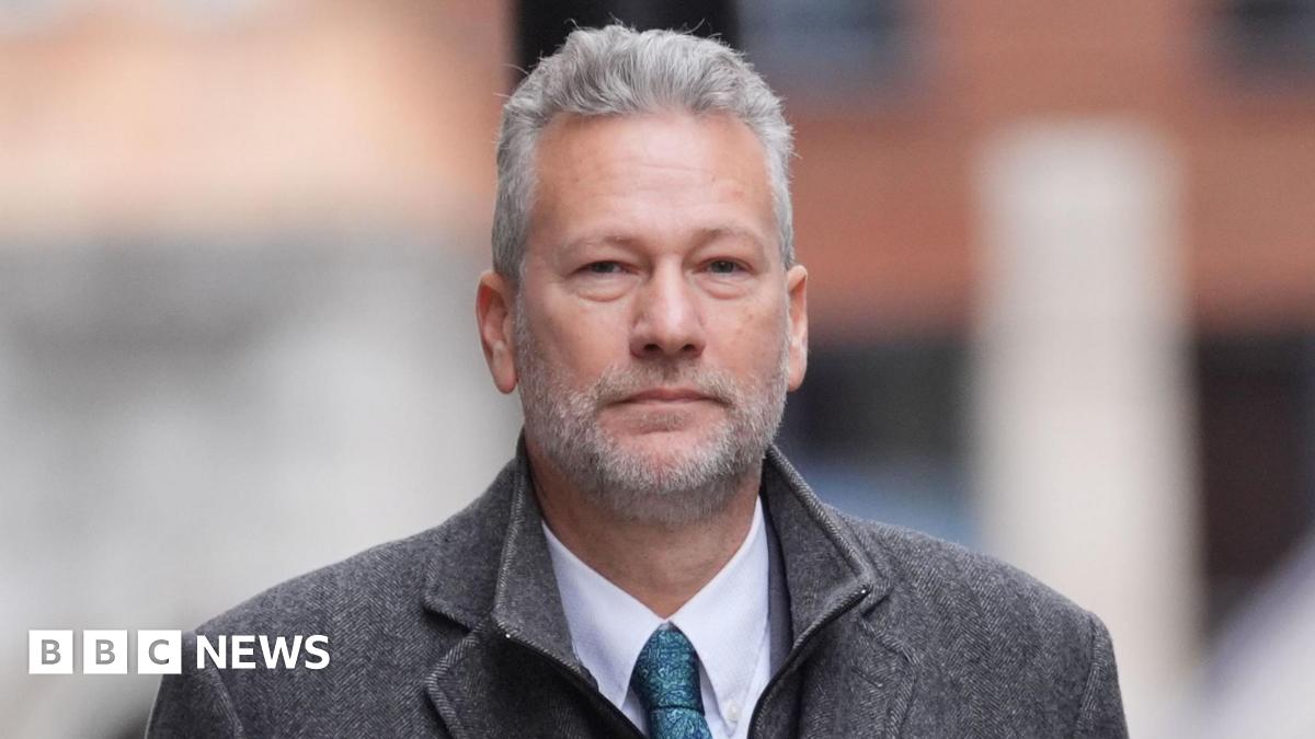 A grey-heaired man with a grey beard wearing a grey coat, blue shirt and a blue tie looking at the camera while walking away from the Old Bailey