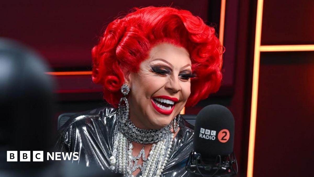 Paul Costelloe has receding grey hair. He is wearing a bright pink, blue and green scarf and a blue jacket.