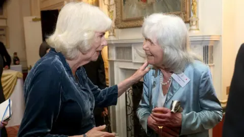 PA Media Queen Camilla and Dame Jilly Cooper at a reception at Clarence House. Dame Jilly wears a pale blue silk suit, while the Queen wears a dark blue velvet jacket. A grand room with lots of other people is visible in the background. 