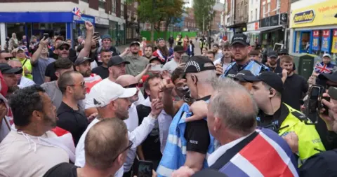 PA Media A large group of people with some holding the flag of England and union jacks, stand very close to a number of police officers who are patrolling the protest in Nuneaton.