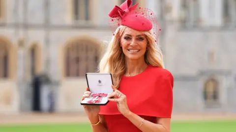 PA Tess Daly holds her medal after being made a Member of the Order of the British Empire (MBE) at an investiture ceremony at Windsor Castle. She wears a red dress and a fascinator. 