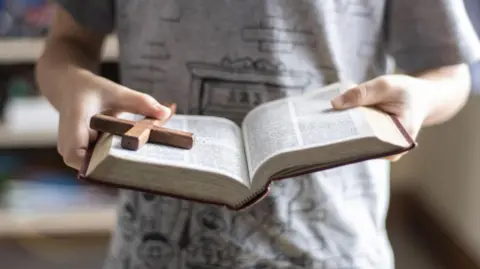 Getty Images A person holding an open Bible with a cross on top of it. The picture is cropped to just show the book in a person's hands. The person is wearing a grey top.