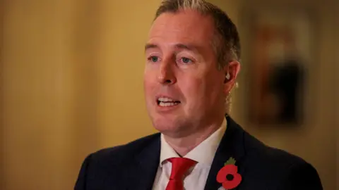 Liam McBurney/PA Wire A photo of Paul Givan mid-speech at Stormont.  He has short, greying hair and is wearing a black suit jacket, a white shirt, a bright red tie and a red poppy on hs lapel. 
