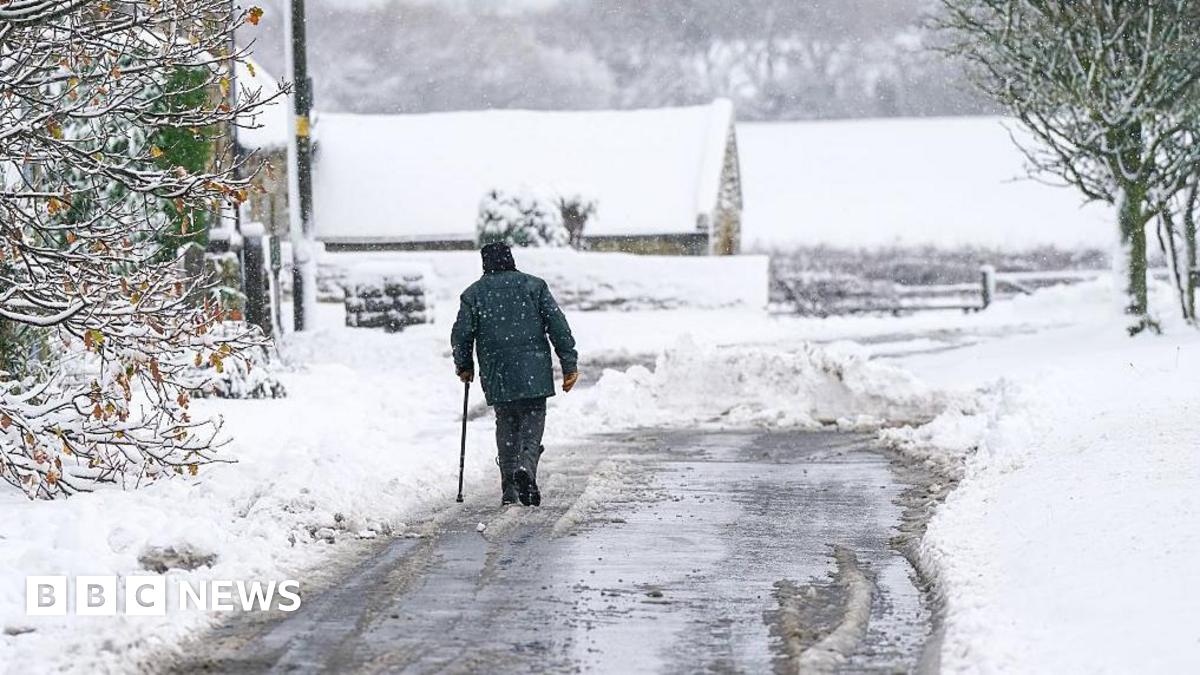 Man walks on a road partially cleared of snow, with a covering of snow on trees in the foreground and a barn and fields in the background
