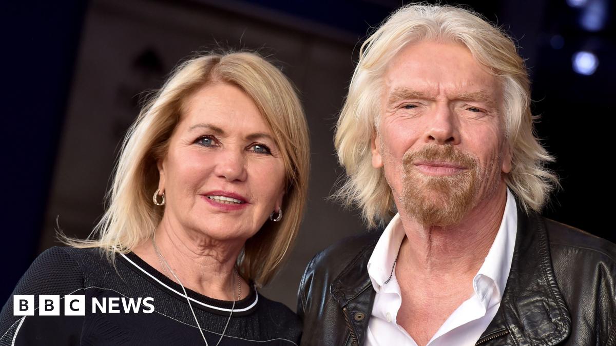 Joan Templeman and Sir Richard Branson look towards the camera. She has long blonde hair and a dark top. Branson has light long hair, a beard, white shirt and brown leather jacket.