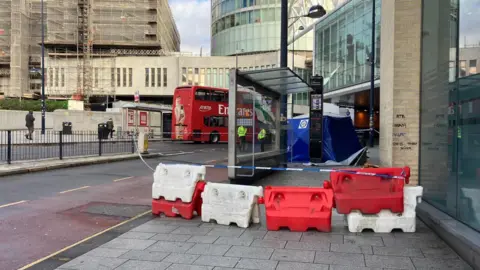 red and white blocks are blocking off a pavement and there is blue and white police tape. a silver bus stop is behind the tape and there is a blue police tent on the pavement beyond it