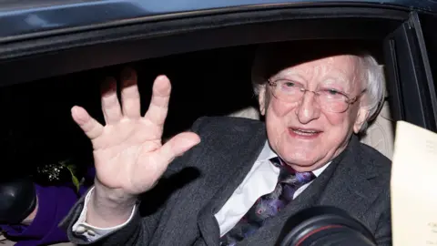 PA Media Michael D Higgins has white hair and glasses and is sitting in a car, waving.