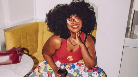 Stephanie Yeboah A woman wearing her natural afro hair is smiling at the camera. She's sitting on a yellow corner armchair, slightly leaned forward. She is wearing funky 70s inspired tinted glasses, a red top, and denim trousers with lots of little animals and fruits on!