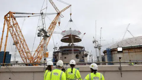 Getty Images A media crew gathers to witness the world's largest land crane 'Big Carl' lifting a 245-tonne domed roof onto Hinkley Point C's reactor building in the background of the image