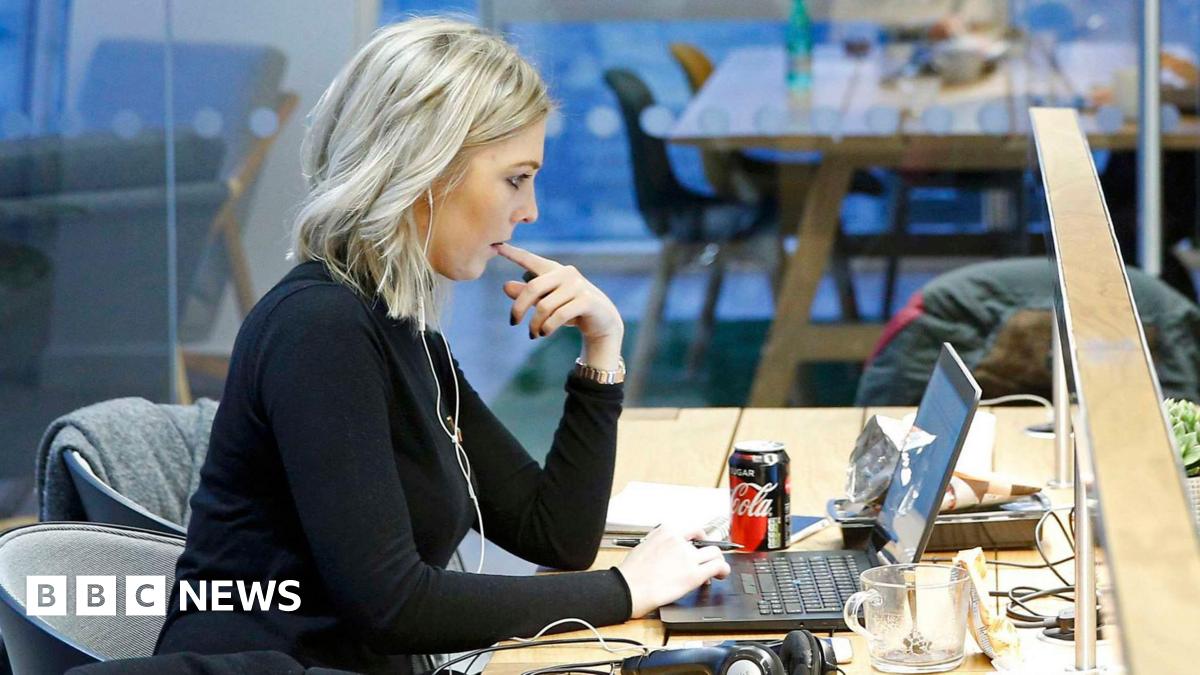Woman with blonde hair sitting at a desk in front of a laptop in a well- lit office. She is wearing a black top and a pair of white in-ear headphones in her ears. She is holding her left index finger in her mouth while using the trackpad on the laptop keyboard.