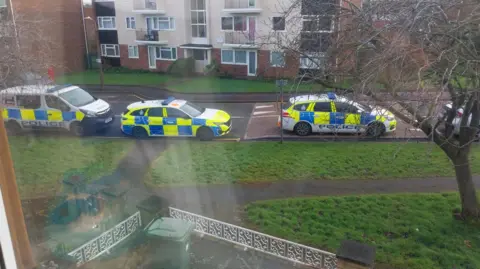 Supplied Seen through an upstairs window, two police cars and a van parked on the street next to a grass verge and opposite a block of flats.