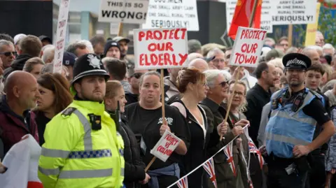 BBC / Claudia Sermbezis People standing with placards behind a Union Jack bunting. There are two police officers standing next to them.