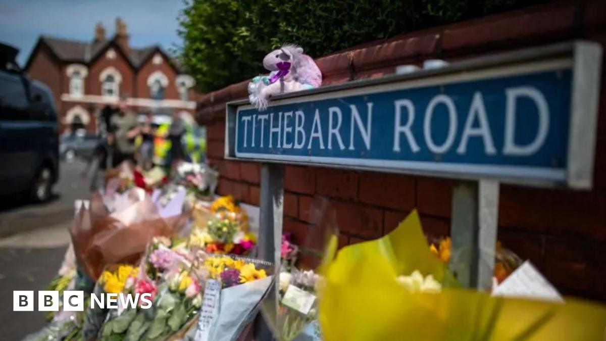 Bunches of flowers and teddies lined up against a wall next to a road sign reading Tithebarn Road.