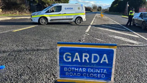 BBC A blue Garda road closed sign sits in the middle of a road. In the background, there's a white van with Garda written on it and an officer is speaking to the driver of a grey car. It is a bright day with blue sky off in the distance.