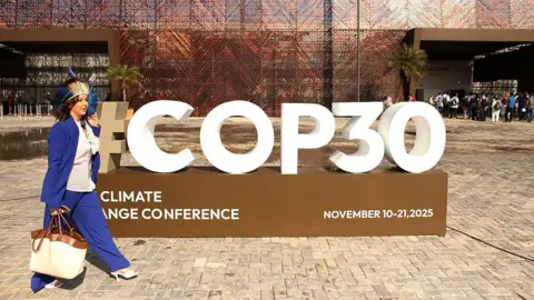 Getty Images A woman in a blue suit and traditional headress, carrying a large white handbag, walks past a large three-dimensional sign advertising "#COP30 climate change conference"