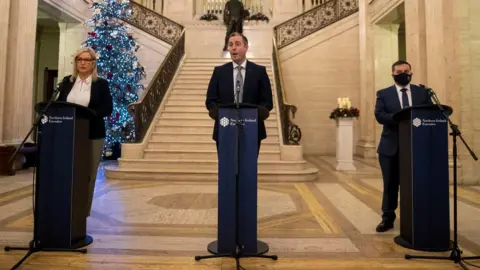 PA Media Michelle O'Neill, Paul Givan and Robin Swann standing behind lecterns in Stormont. A Christmas tree is behind them.