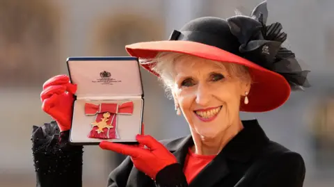 PA Lady Anita May (Anita Dobson) displays her medal after receiving the Officer of the Order of the British Empire (OBE) at an investiture ceremony at Windsor Castle. She smiles straight at the camera and wears red gloves, a red brimmed hat and a black coat. 