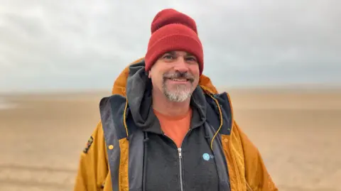 Sam Dixon-French/BBC Dominic Manning stands on Camber Sands beach. He wears an orange t-shirt, a grey hoody and a yellow waterproof.