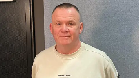 Peter Toole standing next to a black door. He has short grey hair and blue eyes. He is wearing a cream long-sleeved top with a small black writing on it.