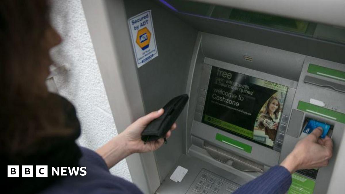 Close-up of a woman using an outdoor ATM machine. The person is holding a wallet in one hand and inserting a blue bank card with the other. The ATM is grey with green accents around the card slot and keypad and the screen displays a welcome message. A security sticker reading ‘Secured by ADT’ is visible on the side panel.