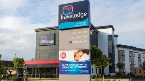 Getty Images The outside of a Travelodge hotel with a nearby sign promising customers will "sleep soundly with great value rooms" beneath a picture of a guest smiling in bed.