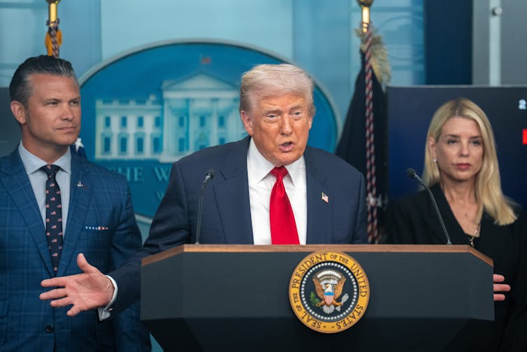 A man in a blue jacket, white shirt and red tie at a lectern, speaking.
