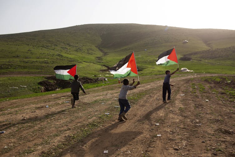 Three boys run down a dirt road, each carrying a Palestinian flag