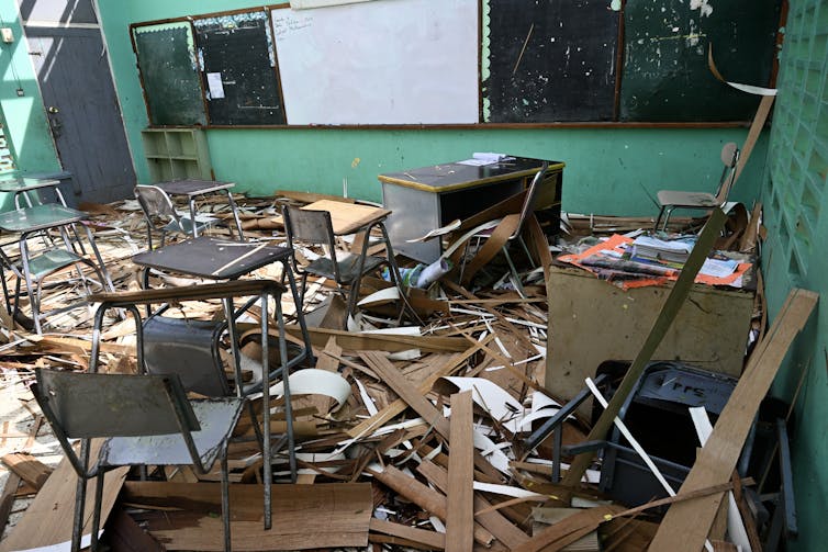 The interior of a school that has been torn apart by hurricane winds. Desks and debris are scattered and light shines through the rafters