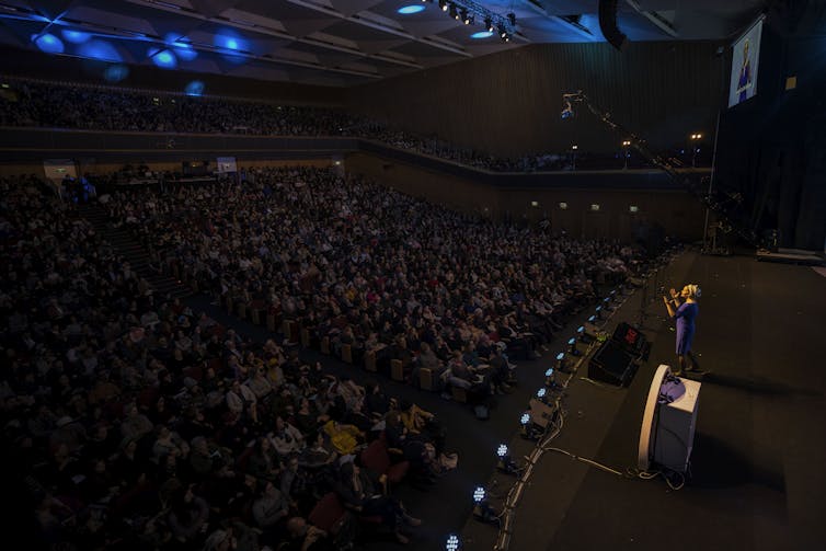 A woman in a purple dress and headwrap gestures as she speaks on stage, facing a large, darkened auditorium full of people.