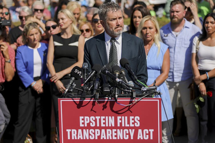 A man in a suit with a crowd behind him stands at a microphone-covered lectern that has a sign 'EPSTEIN FILES TRANSPARENCY ACT' written on it.