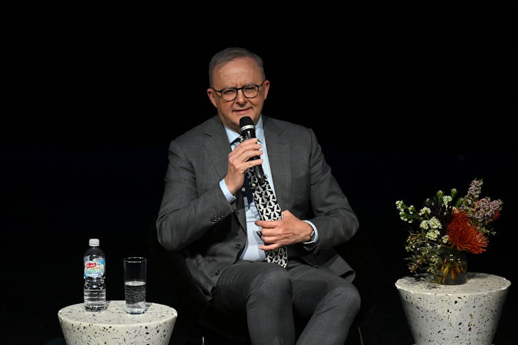 Anthony Albanese sits between two stools on a stage and speaks into a microphone