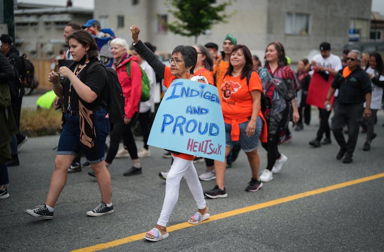 People walk carrying signs and one woman lifts her arm while carrying a sign that says 'Indigenous and proud.'