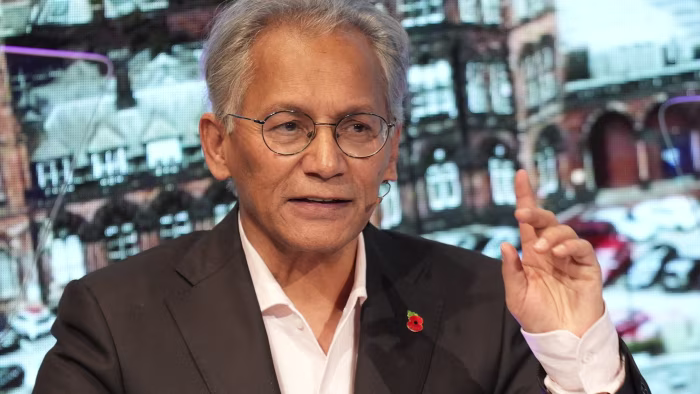 Samir Shah gestures while delivering a speech, wearing a suit and glasses, with a microphone headset and poppy pin.