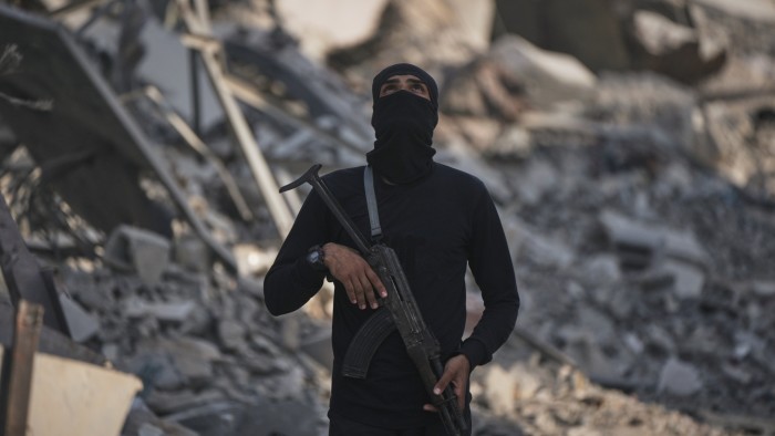 A masked Hamas militant holding a rifle stands amid rubble in Shijaiyah, Gaza City.