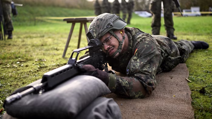 A Bundeswehr recruit in camouflage uniform and helmet lies prone, aiming a G36 assault rifle during basic training.