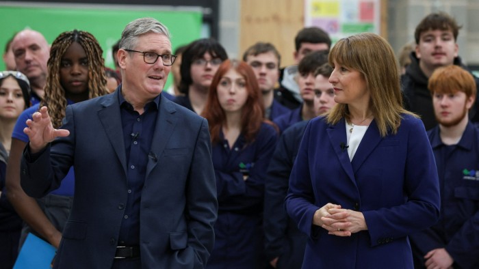 Sir Keir Starmer gestures while speaking beside Rachel Reeves, with a group of students and staff watching in the background.